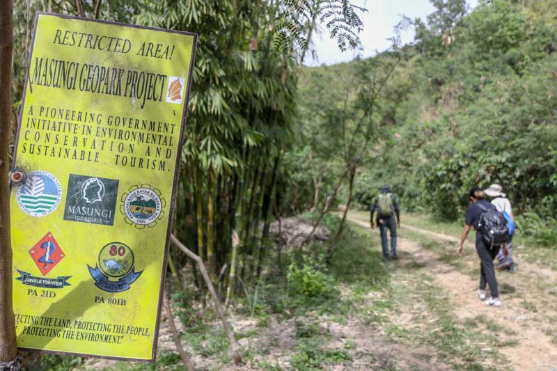 To the left, a sign hangs from a tree indicating the Masungi Geopark Project is a restricted area. To the right, three people wearing hiking clothes walk along a path.